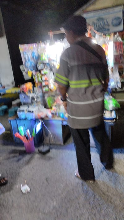 In Sukodono, Indonesia, at night, a person stands facing away from the camera, wearing a gray t-shirt with horizontal green and white stripes and black pants. They are positioned in front of a stall displaying various items, including what appear to be bottled liquids, toys, and other merchandise. The stall is illuminated by bright lights, casting a glow on the surrounding area. The ground is paved, and there is a pink bucket-like container to the left of the stall. A piece of trash is visible on the ground in the lower-middle section of the image.