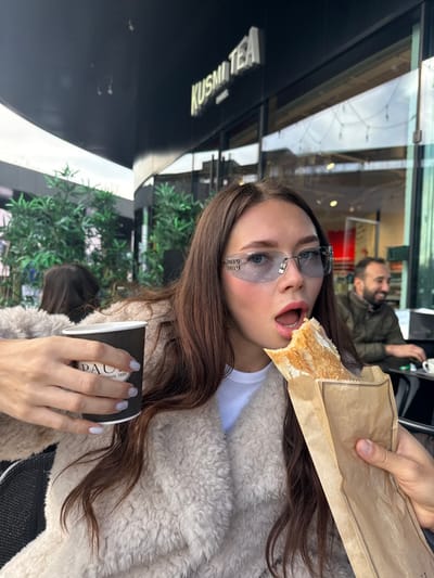 An outdoor scene in Villefontaine, France, shows a young woman with long brown hair, wearing a light beige textured coat and light blue tinted rimless sunglasses bearing "MIU MIU" branding on the temples. She has pink lipstick applied and her mouth is open wide, actively taking a bite from a baguette sandwich. The baguette, containing visible filling, is held in a brown paper bag by a male hand. In her left hand, she holds a black disposable cup printed with "PAUL Depuis 1889". Behind her, a man with a beard is visible, seated at a table, smiling and wearing a dark green jacket. The background features a modern building with a dark, curved facade. A prominent white illuminated sign on the building reads "KUSMI TEA PARIS". Green potted plants are positioned around the outdoor seating area.