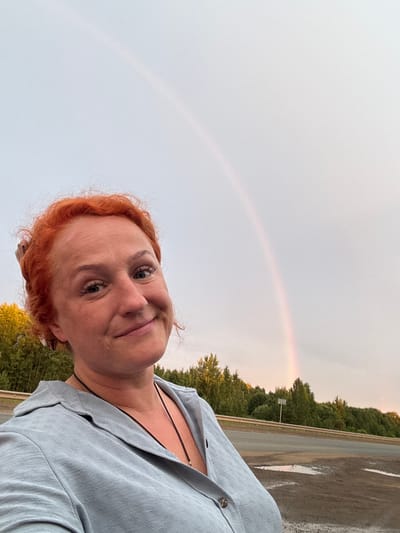 This is a portrait of a woman with red hair taken in Sovkhoznyi, Russia. She is smiling and looking at the camera. She is wearing a light blue shirt and a black necklace. In the background, there is a rainbow and a road with trees. The sky is overcast, and it appears to be late afternoon or early evening. The overall mood of the image is cheerful and peaceful. There is no visible text in the image.