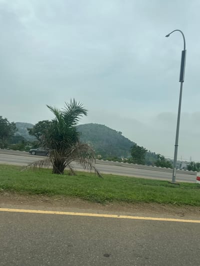 This media file captures a landscape scene, likely in Kunkwaba, Nigeria, on an overcast day. The foreground is dominated by a paved road with a yellow dividing line, suggesting a highway or main thoroughfare. Adjacent to the road is a strip of green grass.
A notable object in the scene is a street lamp with a curved arm, its light fixture appearing to be a solar panel. Further into the scene, a palm tree with dry fronds stands next to a dark-colored vehicle parked on the side of the road.
In the background, a lush green hill or mountain rises, partially obscured by mist or clouds, indicating a potentially humid or rainy atmosphere. Trees dot the slopes of the hill. The overall impression is one of a natural landscape adjacent to a man-made road, with the weather contributing to a subdued and somewhat hazy visual. There is no visible text in the image.