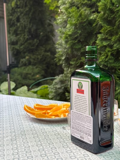 In the city of Pribrezhnyi, Russia, a bottle of Jägermeister sits on a table with a patterned tablecloth. The bottle, a dark green glass with a distinctive embossed logo, is positioned to the right of the frame. To the left of the bottle, a plate holds several slices of orange. In the background, lush green trees create a natural, outdoor setting. The lighting suggests it is daytime, and the overall atmosphere is relaxed and casual.