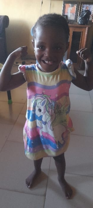 A young Black child stands centrally in a room in Ashipa, Nigeria, facing the camera directly. The child, with short dark hair and small earrings, smiles broadly, revealing teeth, and holds both arms bent at the elbows with clenched fists raised, mimicking a "strong" pose. She wears a sleeveless, knee-length dress patterned with horizontal pastel stripes (yellow, pink, purple, blue) and a large white unicorn graphic on the front. The child is barefoot on a light-colored tiled floor. In the background, a plain, light-colored wall is visible. To the right, a dark wooden cabinet holds at least two framed pictures. To the left, a portion of dark furniture, possibly a sofa or chair, is visible.