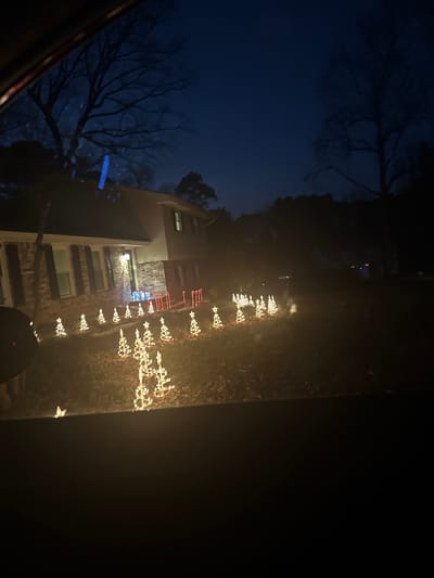 An evening scene shows a building decorated with Christmas lights. The multi-story structure features red brick and white siding. Strings of lights in the shape of Christmas trees and stars line the lawn. Bare trees are visible against a dark blue sky. The perspective suggests the photo was taken from inside a vehicle. The right side of the image is dark, possibly due to being inside the vehicle.