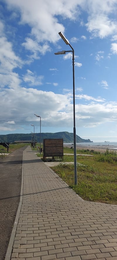 The image depicts a paved walkway with a brick pattern leading alongside a grassy area towards a body of water, likely the Sea of Okhotsk. In the background, a verdant, tree-covered hill slopes down to the coastline. The sky is blue with scattered white clouds, and a large, monolithic street lamp stands prominently on the right side of the walkway. Further down the path, several other street lamps are visible, as well as a wooden structure with a slatted facade, possibly a changing room or information booth. The scene appears to be a public area in Aleksandrovsk-Sakhalinskiy, Russia, possibly a coastal promenade or park, with no people directly visible in the frame. The lighting suggests it is daytime, likely late morning or early afternoon.