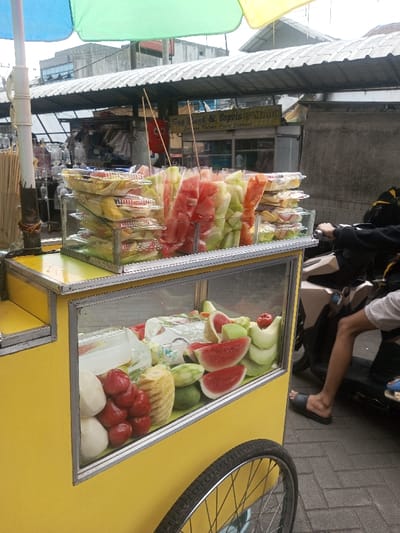 A yellow cart laden with assorted fruits sits on a brick sidewalk. Slices of watermelon, honeydew melon, jicama, pineapple, and rose apples are visible through a glass display case. Above, pre-packaged portions of fruit salad on sticks are stacked high under a colorful umbrella. A person on a scooter appears ready to purchase the fruit. A storefront is in the background.