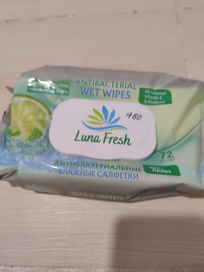 The image shows a package of Luna Fresh antibacterial wet wipes with a lime scent. The packaging features an illustration of a lime slice and blue and green floral graphics. Text on the package includes "ANTIBACTERIAL WET WIPES", "Ph balanced Vitamin E D-Panthenol", "Luna Fresh", "460", "72 pcs/шт", and "Аромат ЛАЙМ" (Aroma LIME). The lower portion of the package displays Russian text: "АНТИБАКТЕРИАЛЬНЫЕ ВЛАЖНЫЕ САЛФЕТКИ" (ANTIBACTERIAL WET WIPES). The product is resting on a light-colored, textured surface, possibly a wooden table. The image itself is somewhat blurry, suggesting it was taken without professional equipment or in low light conditions. There are no people or specific city locations visible in the image.