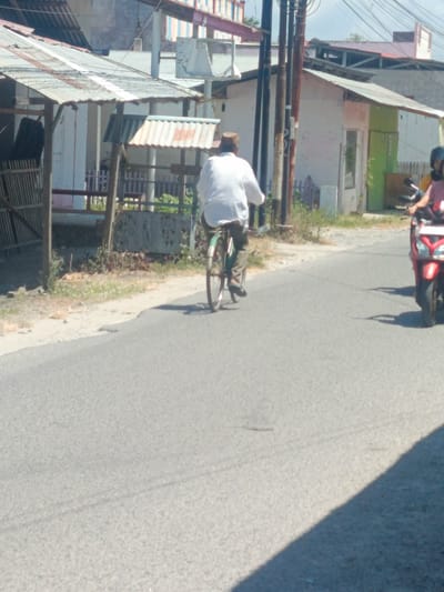 In the city of Tenggela, Indonesia, a man wearing a white shirt and a brown hat rides a bicycle on an asphalt road. The man is in motion, with the bicycle's wheels visible. To the right of the cyclist, a red motorcycle is parked or moving, with its rider partially visible. The background consists of buildings with corrugated metal roofs and some greenery. The scene appears to be a street with typical urban infrastructure, suggesting a common day in the locality.