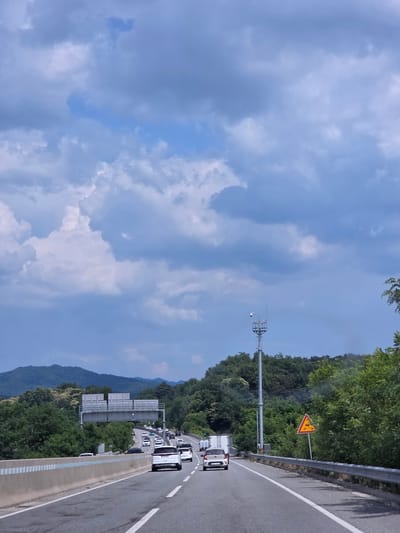 The image shows a view from the driver's seat of a vehicle traveling on a highway in Cheongju-si, South Korea. The highway is a multi-lane road with concrete barriers separating the lanes and the shoulder. The weather is partly cloudy with large, dramatic clouds in the sky. There is moderate traffic on the highway, with cars and trucks visible in various lanes. On the right side of the road, there is a yellow triangular warning sign with an image of a fallen tree and a car, indicating a hazard. A tall metal pole with a camera and antenna is also visible on the right side of the highway. The surrounding environment consists of lush green trees and rolling hills in the background, suggesting a rural or semi-rural setting. Overhead signs with Korean text are visible in the distance, indicating upcoming highway exits or information. The overall impression is of a typical highway drive on a day with dynamic cloud formations.
