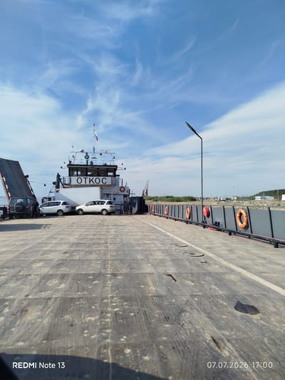 A wide-angle, eye-level outdoor shot shows a ferry docked in Nizhny Bestyakh, Russia. The ferry, named "OTKOC" in large black letters, is positioned in the mid-ground, its ramp lowered. Two white cars and a dark SUV are parked on the ferry deck. The deck itself is made of weathered, grey material with tire tracks and some debris. In the background, a few people stand near the SUV. The sky is a bright blue with scattered white clouds. A tall, dark lamppost with multiple lights stands on the right side of the ferry. The railing of the ferry is dark grey, with several orange life preservers attached. In the far background, there is a coastline with vegetation, industrial structures, and cranes. The date and time "07.07.2026 17:00" and the text "REDMI Note 13" are visible in the bottom left and right corners of the image.