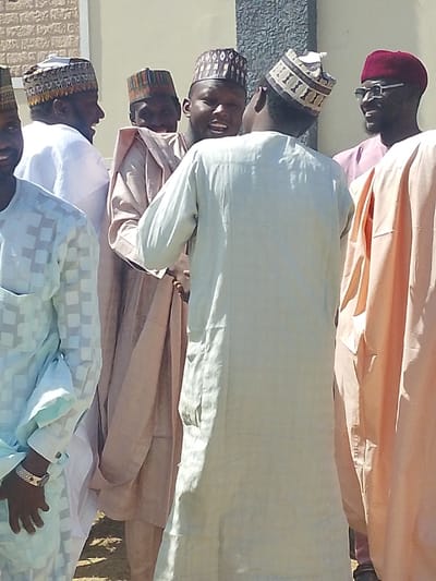 Six adult males are observed gathered outdoors in Darmanawa, Nigeria, under bright daylight. The background is an off-white, textured building wall, possibly stucco or brick. The subjects are dressed in traditional Nigerian attire, consisting of long robes and embroidered caps. From left to right: A partially visible subject wears a light blue, checkerboard-patterned robe and a patterned cap, with a silver-toned wrist ornament on his left wrist. The next subject wears a white robe and a dark patterned cap, smiling towards the right. Another subject in a light brown robe and dark patterned cap smiles towards the right. The central figure stands with his back to the viewer, wearing a plain light green long robe and a light-colored patterned cap. To his right, a partially visible subject wears a light brown robe. On the far right, a partially visible subject wears a reddish-purple cap and eyeglasses, smiling towards the left. The visible subjects exhibit smiling expressions, indicating a social interaction. The ground beneath them is indistinct soil.