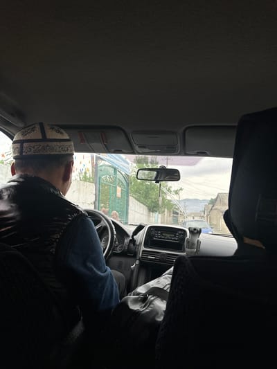The image shows the interior of a car, viewed from the back seat. The driver, a man wearing a traditional Kyrgyz hat and a dark vest over a blue shirt, is seen from behind, looking forward and gripping the steering wheel. The dashboard of the car is visible, with a radio and a phone mount. Outside the car, through the windshield and side windows, a street scene in Nookat, Kyrgyzstan, is depicted. A green gate and a wall are prominent on the left side, with trees and buildings in the background. The sky is overcast, suggesting it might be a cloudy day.