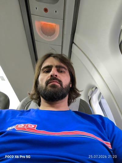 The image shows a man with brown hair and a beard taking a selfie on an airplane. He is wearing a blue t-shirt with a red and white stripe design. The airplane cabin is visible in the background, including an overhead light and air vent. A timestamp in the lower right corner indicates the photo was taken on July 25, 2026, at 21:20. The bottom left corner shows "POCO X6 Pro 5G". There is no information in the image that suggests this location is in Kogalym, Russia.