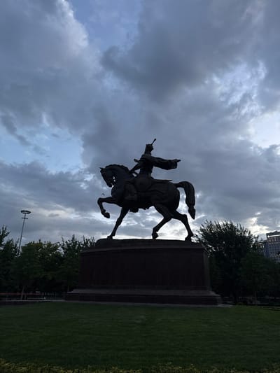 The image shows a statue of a man on horseback silhouetted against a cloudy sky. The statue stands on a large, multi-tiered pedestal, surrounded by greenery. A building is visible in the background. Based on the location information, it is likely located in Kashgar mahalla, Uzbekistan.