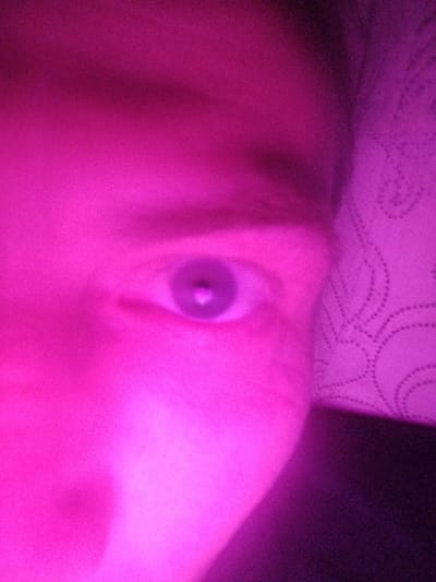 The image shows a close-up of a person's face, specifically the area around one eye. The skin and surrounding area are heavily tinted with a bright magenta color, possibly due to lighting. The eye is visible, with the pupil and iris discernible. Part of a patterned surface, likely fabric, is visible to the right of the face. The image lacks contextual details to provide a location in Gus-Khrustalny, Russia or to describe actions or interactions.