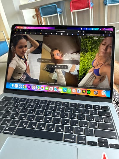 An overhead medium shot shows a laptop with a video call displayed on its screen. The laptop is positioned on a person's lap, who is wearing patterned shorts. The video call features three different feeds. The feed on the left shows a young woman with dark hair, wearing a white collared shirt, with her hand on her head. The central feed shows a woman with blonde hair, wearing a blue bikini top, with the text "hi." overlaid on the screen. The rightmost feed also shows a woman with blonde hair, wearing a blue bikini top, with a lush green background. The bottom of the laptop screen displays a dock with various app icons. The laptop is placed on a surface, possibly a table, with a blurred background showing chairs and a window. The overall scene appears to be indoors. There is no visible indication that the image was taken in Sao Thong Hin Town Municipality, Thailand.
