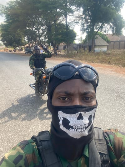 The image depicts two individuals on a paved road in Sankara, Nigeria, under clear daylight conditions. In the foreground, a dark-skinned individual takes a selfie, partially obscuring their face with a black balaclava featuring a white skull design, and wearing black-framed goggles pushed onto their forehead. They are dressed in a camouflage military uniform with a tactical vest. Immediately behind this individual, a second dark-skinned person rides a dark-colored motorcycle. This person is also in a camouflage military uniform, wearing dark sunglasses, and is smiling while gesturing with their right hand in a "V" sign.
The environment includes a two-lane asphalt road with visible cracks. To the right of the road, the ground is dry and dusty with sparse vegetation. The background features several mature trees with green foliage. Beyond the trees and road verges, multiple single-story buildings are visible, some with corrugated metal roofs and light-colored walls. One building displays a blue and white striped pattern on its lower section. A light-colored pickup truck is partially visible parked further down the road on the left side. Shadows indicate strong sunlight from the right side of the frame relative to the foreground subject.