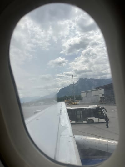 The image is taken from an airplane window, looking out at an airport tarmac. In the foreground, the wing of the airplane is visible, along with part of an engine. A white and black bus, labeled "COBUS 3000," is parked on the tarmac. A person in uniform, appearing to be security personnel, stands beside the bus. In the background, mountains are visible under a cloudy sky. The airport appears to be situated in a valley, possibly Viehhausen, Austria, given the proximity to the Alps. There are buildings, possibly hangars or terminals, and various airport vehicles scattered across the tarmac. The overall scene depicts a moment on the ground before or after a flight, with a backdrop of impressive natural scenery.