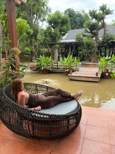 A woman wearing a leopard print jumpsuit and tan sneakers is reclining on a large, round, dark wicker chair with a gray cushion. She is facing away from the camera, looking towards a pond with a small fountain in the center. The chair is situated on an orange tiled patio. Behind the patio is lush greenery, including trees and large leafy plants. In the background, there is a building with a dark roof and what appears to be a wooden deck extending over the pond. The location is identified as Mae Rim, Thailand.