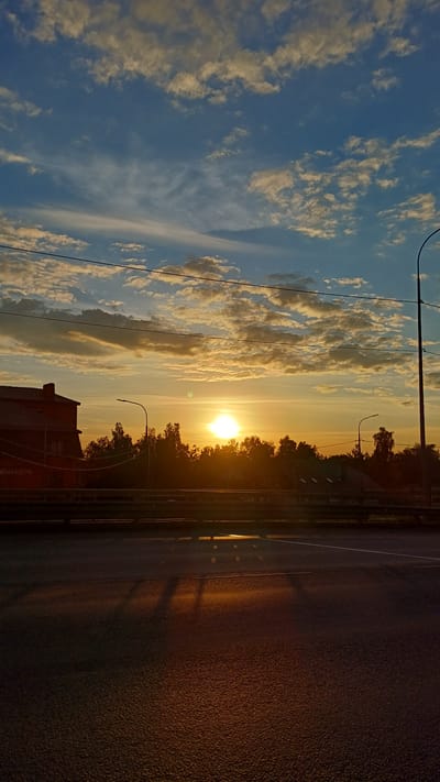 The image captures a serene sunrise or sunset scene, viewed from the side of a road. The sky dominates the upper portion, displaying a clear blue transitioning into warm golden and orange hues near the horizon. It is adorned with numerous fluffy and elongated clouds, brilliantly illuminated by the low sun.
A bright, intense sun is prominently positioned near the horizon, partially obscured by a dense band of silhouetted trees and foliage. This creates a dramatic contrast against the glowing sky and casts warm light across the entire landscape.
In the foreground and extending along the right side, a dark asphalt road surface is visible, textured and reflecting a warm, subtle glow from the sunlight. Long, dark shadows, possibly from overhead utility poles or other structures, stretch diagonally across the road. Several slender utility poles or streetlights with overhead wires are silhouetted against the bright sky, extending upwards from the ground.
On the bottom left, a section of a reddish-brown building with visible windows is partially captured. Faint Cyrillic text, "СЕРВИС АВТО" (meaning "Service Auto"), is discernible on its side. The overall lighting indicates either early morning or late evening, imbuing the scene with a peaceful and atmospheric mood. No people or specific activities are visible within the frame.