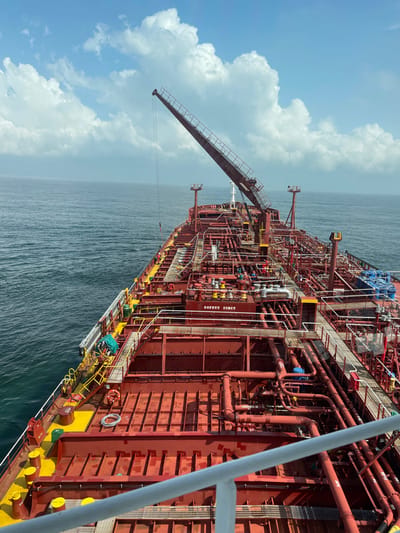 The image displays the forward deck of a large, red-hulled vessel, appearing to be an oil tanker, underway at sea off the coast of Orimedu, Nigeria. The sky is clear blue with scattered white cumulus clouds, and the ocean is calm, extending to the horizon.
The ship's deck is predominantly deep red, featuring an intricate network of interconnected red pipelines of varying diameters, equipped with numerous valves and fittings. Yellow-painted walking paths and mooring bollards provide contrast. A dark-colored crane boom extends diagonally upwards and forward from the midship area, with a cable suspended from its tip. A white sign, prominently displaying "SAFETY FIRST" in red text, is affixed to a central deck structure.
On the port side, an individual wearing a blue or green protective suit is visible near a yellow-railed gangway, engaged in an unspecified activity near the ship's railing. Coiled white ropes or hoses are present on the deck in this vicinity. On the starboard side, midship, several blue barrels or containers are stacked. White railings delineate elevated platforms and the ship's perimeter. The vessel is in open water, with no land visible beyond the horizon.
