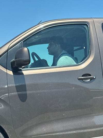 A man wearing glasses and a light blue t-shirt is driving a dark-colored vehicle. The man is viewed in profile from the side, facing right. He is holding the steering wheel with both hands. The vehicle's windows are tinted, obscuring the interior beyond the driver. The exterior of the vehicle appears dusty. The background is a clear blue sky, suggesting an outdoor setting. The image was taken in Palauzovo, Bulgaria.