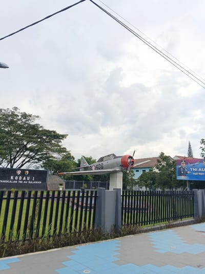A gray and red airplane, marked as "BO 408", is mounted on a white pedestal behind a black fence. Behind the fence, green grass is visible. A black sign on the left displays white text. A poster is on the right, with a military figure wearing a uniform. Trees and buildings are visible in the background under a cloudy sky, and power lines stretch across the upper portion of the image. A tiled walkway is in the foreground.