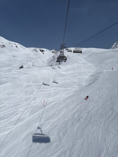 The image displays a snow-covered alpine ski slope in the Sonnenkopf ski area near Klösterle, Austria, under a clear blue sky. A chairlift system, identifiable in part by "PLANIWOS" on one carrier, extends across the scene. Its steel cables run diagonally through the upper and middle sections of the frame. Multiple chairlift chairs are visible along the cable, with shadows of several chairs, including the one from which the photograph appears to be taken, cast sharply onto the snow. At least two individuals are seated in one of the ascending chairs. In the lower-right quadrant, a solitary skier wearing a red jacket and dark pants is actively descending the slope. The snow is extensively marked with ski tracks from previous activity. Several red marker poles are positioned on the slope, and a small cluster of evergreen trees is visible on a snowfield to the left. Distant mountain peaks show areas of exposed rock.