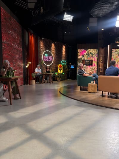 The image depicts a television studio set in Sant Joan Despí, Spain, likely for a show called "La Selva." The set features a brick wall backdrop with a circular neon sign that reads "LA SELVA." In the foreground, several people are seated at tables and on chairs, appearing to be guests or participants in a broadcast. One woman in a light-colored top and green pants sits at a table on the left, with a glass of water in front of her. Further back, a man in a hat and a woman in a dark top are seated at another table. To the right, a man in a grey t-shirt and a woman in blue jeans are seated on a green sofa. Another person in a blue jacket is visible in the background. The studio is equipped with lighting rigs and decorative elements such as plants and a vintage-style jukebox. The overall atmosphere suggests a casual and engaging television program.
