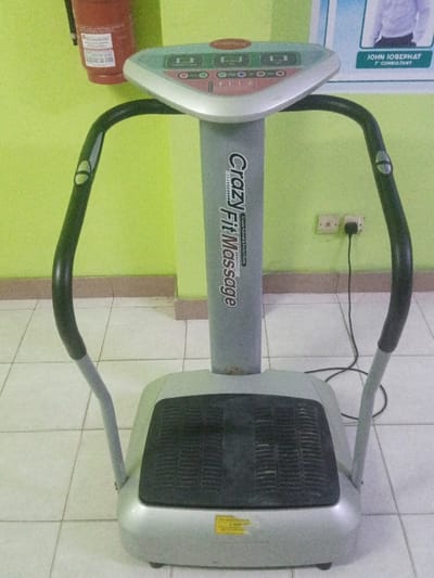 A "Crazy Fit Massage" vibration exercise machine is centrally positioned in a room in Majengo ward, Kenya. The machine is predominantly silver-grey, featuring a black textured foot platform and two curved black handlebars. Its central vertical column displays "Crazy Fit Massage," and "CrazyFit" is visible on the top control panel, which includes an array of buttons and indicator lights. A black power cord extends from the machine's base, plugging into the right socket of a white two-gang electrical outlet mounted on the right wall. The room's walls are painted a vibrant lime green. In the upper left background, a red, wall-mounted fire extinguisher is partially visible. To the upper right, a portion of a poster displaying a person in a white shirt and the text "JOHN JOSEFAT T CONSULTANT" is present. The floor is covered with white square ceramic tiles separated from the green wall by a light brown baseboard. No individuals are visible in the scene. The machine appears functional and shows minor signs of wear and dust.