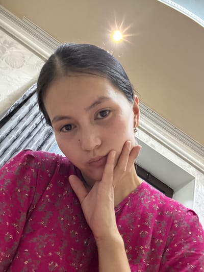 A young woman with dark hair pulled back is taking a selfie indoors. She is wearing a bright pink shirt with a subtle floral pattern. Her left hand is raised to her cheek, and she is looking down at the camera with a pouty expression. The background suggests an interior room with patterned wallpaper, curtains, and architectural molding, possibly in the city of Жыгач-Коргон, Kyrgyzstan. A light fixture is visible on the ceiling.