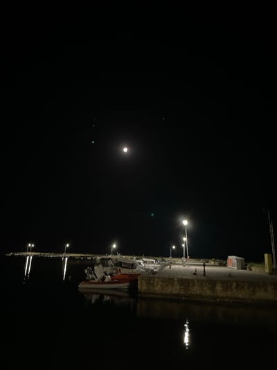The image depicts a nighttime scene at a harbor in Limenas Avdiron, Greece. The sky is dark with a bright full moon visible in the upper center. Several small, faint green lights are also present in the sky. The harbor area is illuminated by streetlights positioned along a stone quay. These lights cast reflections on the dark water. Moored in the water are several boats, including a rigid inflatable boat with an orange hull and a white and gray cabin boat. The quay itself is constructed of stone and concrete and appears to be a public area with some infrastructure like bollards and what looks like a trash receptacle. The overall atmosphere is calm and still, characteristic of a quiet night in a coastal town.