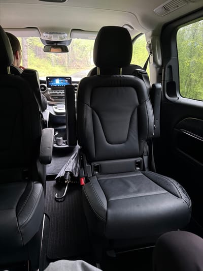 The image displays the luxurious interior of a modern vehicle, likely a van or SUV, from a rear-facing perspective, looking towards the front cabin. The setting is a rainy day in Нареченски бани, Bulgaria.
The primary subjects are the vehicle's interior components and a partially visible person. The cabin is outfitted with black leather captain's chairs, two of which are visible in the foreground, and a front passenger seat. Water droplets are distinctly visible on the upper part of the front passenger seat's backrest and on the vehicle's windows, indicating recent or ongoing rain.
Through the large windows and sunroof, a lush green exterior, presumably a forest or dense vegetation, is visible and appears wet. Inside, a person is resting or sleeping, with their head visible against the front passenger seat's headrest.
Notable objects include a modern digital display (resembling a tablet) on the central console, which appears to show a map or media interface. Below the screen are climate control vents and other dashboard elements, including a dial displaying the number '0'. An umbrella, folded and wet, is placed on the ribbed black floor mat between the rear seats. Overhead, an interior rearview mirror, lighting, and a console with a partially visible "AIRBAG" label are present. The seats also feature "ISOFIX" labels, indicating child seat anchor points, visible on the side of the seats. The vehicle's interior suggests comfort and luxury, providing shelter from the inclement weather outside during what appears to be daytime.