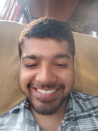 A male subject, appearing to be a young adult with dark hair and a beard, is depicted smiling broadly with his eyes closed or nearly closed. He is wearing a plaid shirt with white and dark checkered patterns. The subject is positioned indoors within the city of Kaliganj, Bangladesh, against a light-brown or beige upholstered background, possibly a seat or wall. Bright sunlight enters from the upper left, illuminating the scene and creating highlights on the subject's hair, with patterned curtains (red and orange hues) partially visible in the light source. A small, circular, caged fan is discernible in the upper right background. A digital timestamp "04/12/2025 14:32" is present in the lower right corner of the image.