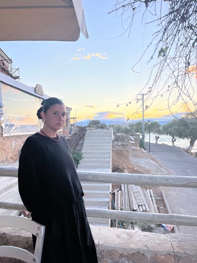 The image depicts an outdoor scene in Megalochori, Greece, during twilight, characterized by a vibrant sunset with hues of orange, pink, and yellow along the horizon fading into a blue sky overhead.

In the left foreground, a female individual, appearing in her 20s or 30s, stands facing the viewer with a neutral expression. She has dark hair styled in a bun, visible hoop earrings, and a dark red beaded necklace. She wears a long-sleeved black top and a long black skirt or dress, leaning her left arm on a light-colored wooden railing that spans the lower portion of the frame. To her immediate left, a partial white outdoor chair is visible. The ground beneath the railing is a rough, cracked concrete or stone surface.

Behind the individual, to the left, is a building undergoing construction or renovation, marked by scaffolding, exposed brick, and transparent plastic sheeting. A small, unreadable wooden sign is affixed to this structure. A white patio umbrella is partially visible in the upper left corner.

Further into the mid-ground, a descending flight of light-colored concrete stairs leads down to the right. Adjacent to the stairs and below the railing, the terrain is uneven earth with construction debris, including piles of rebar and stacked wooden planks.

The background reveals a coastal environment. A paved road or path curves along a body of water, identifiable as the sea, where gentle waves are visible. Trees with green foliage line the road and shore. A black streetlamp with an illuminated globe stands beside the road. A utility pole with power lines is also visible, partially obscured by bare, skeletal tree branches extending into the upper right quadrant of the image. Distant low-lying landforms or mountains are visible on the horizon under the setting sun.