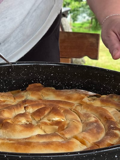 A round, dark-colored baking pan with white speckles contains a freshly baked, golden-brown savory pastry arranged in a prominent spiral pattern, exhibiting a flaky texture with visible white filling. On the left, a person wearing a light grey t-shirt over dark trousers is partially visible, holding a metallic utensil, likely a fork, poised above the pan. On the right, the partial view of a person's right hand and wrist, adorned with a delicate silver-colored beaded bracelet, is present near the pan. The background features a blurred outdoor environment with lush green foliage and a segment of rustic wooden furniture. The scene is located in Gigintsi, Bulgaria.