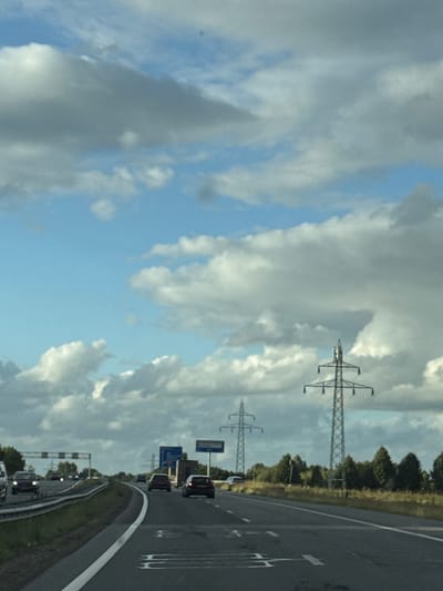 The image shows a view from a car driving on a highway, likely in or near Meppel, Netherlands, given the presence of Dutch road signs. The sky is filled with dramatic, partly cloudy formations. In the foreground, the asphalt road surface is visible with white lane markings. Several cars are driving on the highway in the same direction as the viewer. On the right side of the highway, there are trees and two tall electricity pylons. On the left side, there is a guardrail and more trees. Blue highway signs are also visible in the distance. The overall scene depicts a typical daytime highway commute under a dynamic sky.