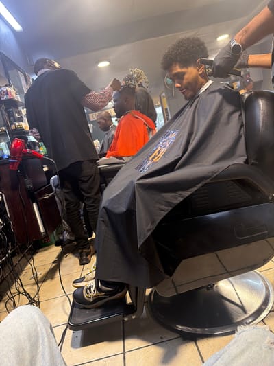 This image captures an active barbershop scene, likely located in Blouberg, South Africa. In the foreground, a barber, wearing black gloves and a striped shirt, is intently trimming the hair of a male client using electric clippers. The client, who has dark curly hair and a short beard, is seated in a black barber chair and covered by a black cape adorned with a distinct, colorful stylized logo. His expression appears calm during the haircut.
The setting features a light-colored tiled floor, where the shiny, circular base of the barber chair is visible, along with several electrical cords running across it. In the midground, another barber in a black shirt and checkered undershirt is attending to a different client with dreadlocks or braids, who is wearing an orange top. This suggests a busy establishment with multiple active stations. A red hairdryer rests on a dark wooden counter in the background. Overhead artificial lighting illuminates the entire scene. The client in the foreground is wearing black and yellow athletic shoes, visible on the footrest of his chair. The atmosphere is that of a functional, professional grooming space.