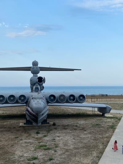 The image depicts a large, gray, winged vehicle, possibly an aircraft or a seaplane, parked on a dry, dusty ground with sparse vegetation. The vehicle has multiple engines mounted on its wings and a prominent cockpit area. In the background, a calm blue sea stretches to the horizon under a pale blue sky with scattered clouds. A sandy beach with a few structures and people is visible between the vehicle and the sea. To the right of the vehicle, a paved walkway runs alongside the scene, and two individuals, one in a red dress and another in a blue garment, are walking away from the camera on the walkway. The setting appears to be an outdoor exhibition or a museum in Arablyar, Russia, possibly a memorial site for the aircraft.