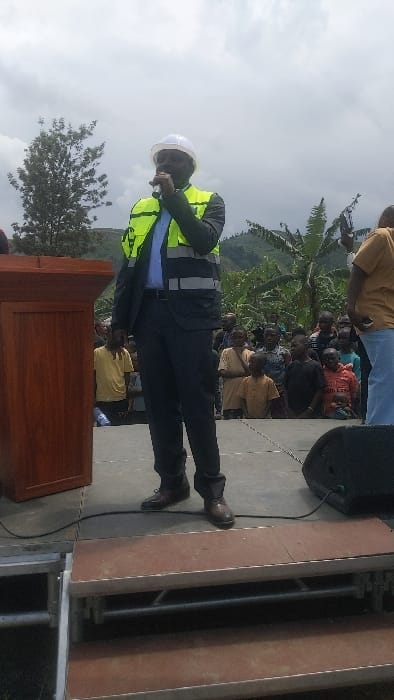 The scene takes place in Sake, Democratic Republic of Congo (latitude: -1.57, longitude: 29.046), with a man in a suit and a white hard hat speaking into a microphone while wearing a bright yellow safety vest. He is standing on a stage with a wooden podium to his left, and several people are standing behind him. The crowd of people listening to him are wearing a variety of clothing, including a yellow shirt, a light blue shirt, a black t-shirt, and a red t-shirt. There is a large speaker on the stage with a black cable running towards it from the left, along with a set of stairs leading up to the stage, built from metal and wood. The background of the image is a lush green landscape with a few banana plants in the distance. The sky is cloudy.
