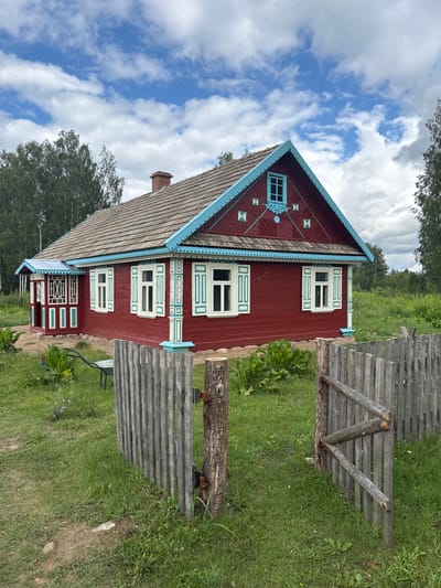 This media file displays a traditional, brightly painted red wooden house with a blue-trimmed roof and decorative window frames, set in a lush, green, grassy landscape. The house features several windows with white shutters and intricate blue and white trim. A weathered wooden fence runs along the side of the property, and a chimney is visible on the roof. The sky is a vibrant blue with scattered white clouds, suggesting a pleasant, possibly sunny day. Trees can be seen in the background, indicating a rural or semi-rural setting. There are no people or animals visible in the image, and no text is present. The scene evokes a sense of rustic charm and tranquility. The orientation of the image is portrait and rotated 90 degrees clockwise.