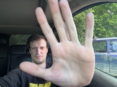 The image shows a man inside a vehicle, holding his hand up to the camera, obscuring his face. The man is wearing a black sweatshirt with a yellow design. The vehicle's interior is visible, including black leather seats. Outside the vehicle, trees and another vehicle are visible. The image was taken on a sunny day.