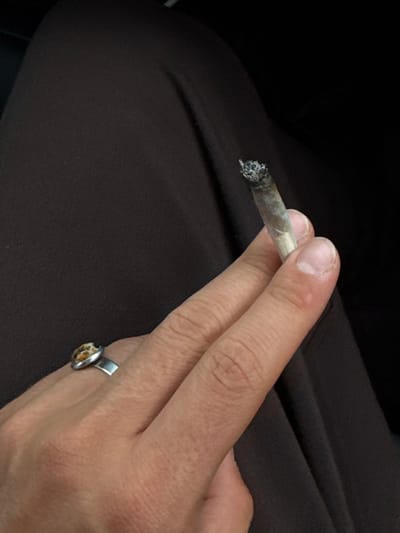 The image provides a close-up, angled view of a human hand holding a lit, partially consumed hand-rolled cigarette or joint. The subject's skin exhibits visible pores and fine lines, with fingernails showing natural cuticles. A silver-toned ring with an oval, mottled yellowish-brown stone is worn on the ring finger. The background consists primarily of dark brown, lightly textured fabric, likely clothing or upholstery, against an even darker, indistinct area, suggesting an indoor or vehicle environment. No discernible details within the image can confirm a specific geographical location in Anzola dell'Emilia, Italy.