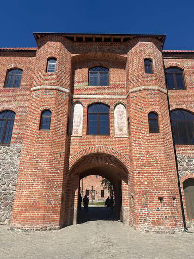 The image shows a brick building in Guryevsk, Russia. The building has several arched windows and an arched entrance. The exterior is constructed primarily of red bricks, with a foundation made of stones. The sky is bright blue. Two silhouetted people are visible passing through the arched entrance. The ground near the building is paved.