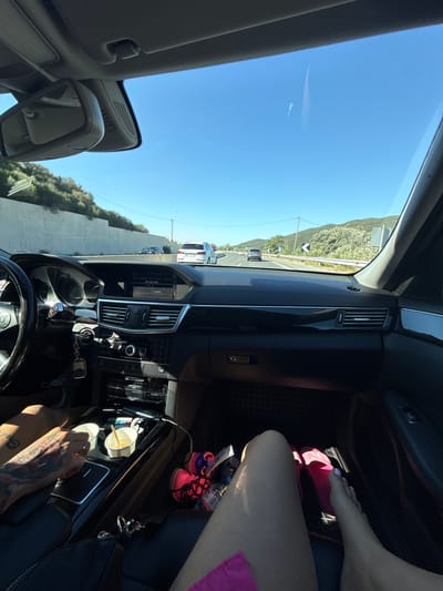 The image shows the interior of a car driving on a road in the Municipal Unit of Promachon, Greece. The view is from the passenger seat, looking towards the driver's side and out the windshield. The dashboard and center console are visible, with a steering wheel on the left and an infotainment screen displaying "FM 104.2 MHZ DARIK". Two paper cups with drinks are in the center console, and a person's tattooed arm is reaching for one. In the back, a person's legs and feet are visible, wearing pink sandals. Outside the car, the road curves through a hilly, green landscape under a clear blue sky. Several other cars are visible on the road ahead, including a white SUV and a darker sedan. The road has a concrete barrier on the left and a guardrail on the right, with signs indicating a curve. The time of day appears to be daytime, with strong sunlight casting shadows.