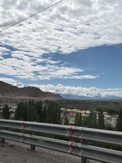 In the city of Phey, India, a view is captured from a slightly elevated position, looking over a valley. In the foreground, a metallic guardrail with white and red striped markings runs horizontally across the frame. Beyond the guardrail, a cluster of tall, slender green trees forms a dense line. Further back, a settlement of buildings with light-colored roofs is visible, nestled amongst more greenery. In the mid-ground, the landscape rises into rolling hills and then a large, imposing mountain range with rocky, barren slopes. The background is dominated by a bright blue sky filled with fluffy white clouds. The overall scene depicts a valley settlement in a mountainous region under a cloudy sky.