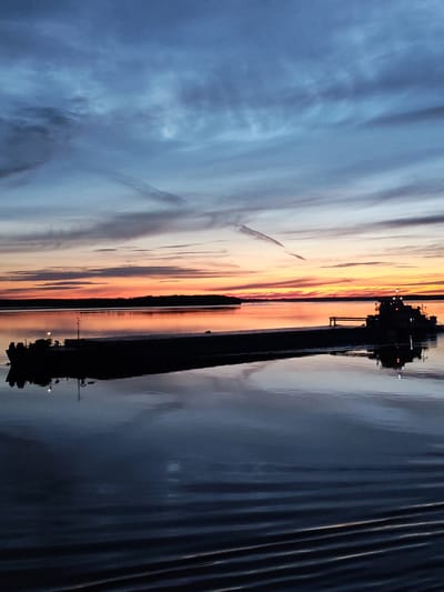 At dusk, a barge with a pilot house is navigating a large body of water. The sky is a gradient of deep blue and orange, with streaks of clouds suggesting an ongoing sunset. The water's surface is calm, reflecting the sky and the barge with a high degree of clarity. A faint contrail is visible in the upper portion of the sky. The scene evokes a serene and tranquil atmosphere. The location is identified as Malinukha, Russia.