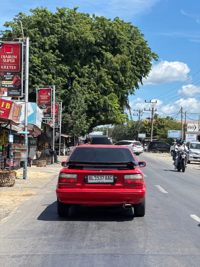 The image shows a street scene in Tumpok, Indonesia. In the foreground, a red car with license plate BL 1537 AAC is driving on a paved road. The car has a spoiler on the back and a visible exhaust pipe. On the left side of the road, there are several shops and stalls, including one with a sign for "Djarum Super Kretek". A large tree with lush green foliage dominates the background, with a bright blue sky and scattered white clouds above. Further down the road, other vehicles, including cars and motorcycles, are visible, as are power lines and utility poles. One motorcycle has two people riding on it. The overall impression is a sunny day in a bustling urban or semi-urban environment.