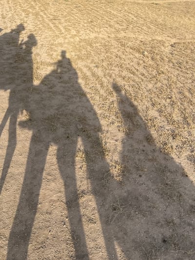 The image depicts a shadow of several people, one holding what appears to be a camera or phone, cast onto a dirt or sandy ground, likely in an arid environment. The ground is covered with sparse vegetation. The scene is possibly located in or near Sidi Badhaj, Morocco.