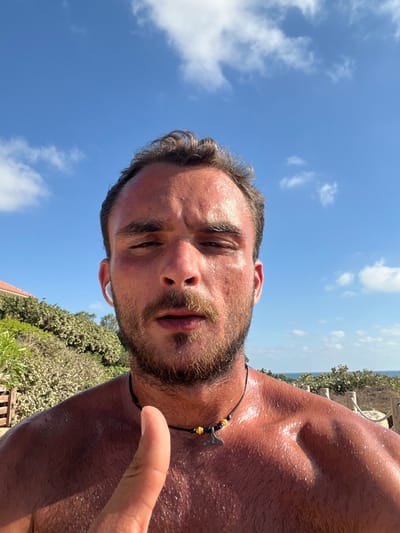 A sun-drenched selfie features a man with a beard, a black necklace with an axe pendant, and sweaty skin under a clear blue sky. He gazes downward, his features highlighted by the strong sunlight. His head is slightly tilted, with one finger pointing upwards. The background shows lush green bushes and a glimpse of blue ocean.