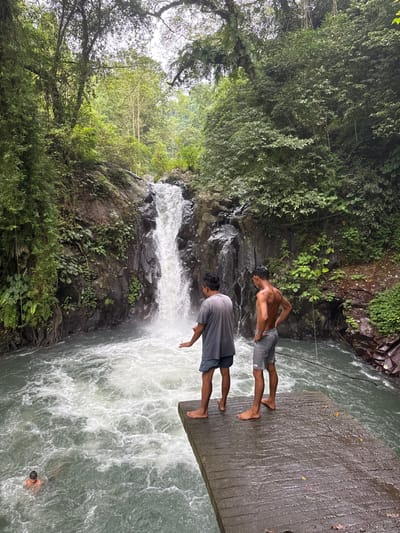 Two men stand at the edge of a concrete platform overlooking a waterfall. The water flows down a rocky cliff surrounded by lush green vegetation. One man is shirtless, while the other wears a grey shirt and shorts. Below, in the turbulent water, a third person is partially submerged, with only their head visible. The scene captures a moment of observation and perhaps adventure at a natural waterfall setting.